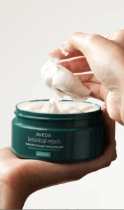 A hand is scooping out a white cream from a green jar labeled "Aveda Botanical Repair Intensive Strengthening Masque Rich.